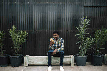 Smiling male worker using mobile phone while sitting on bench against corrugated wall - GRCF00492