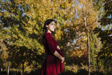 Pregnant woman with hands on stomach standing in park during autumn - EBBF01664