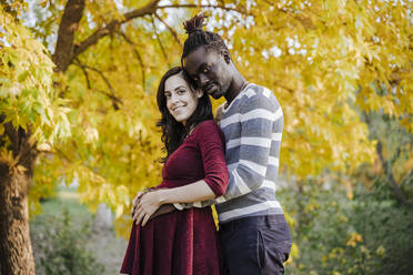 Lächelndes multiethnisches Paar mit Händen auf dem Bauch im Park stehend im Herbst - EBBF01680