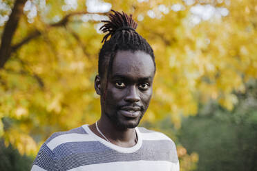 African man standing against yellow tree in park during autumn - EBBF01683