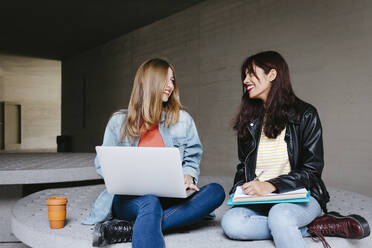 Lächelnde junge Freundinnen, die sich gegenseitig ansehen, während sie mit Büchern und Laptop auf dem Campus sitzen - TCEF01348