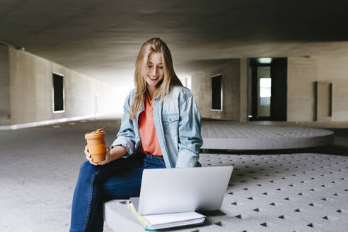 Lächelnde blonde Studentin, die auf einen Laptop schaut, während sie mit einem wiederverwendbaren Kaffeebecher auf dem Campus sitzt - TCEF01352