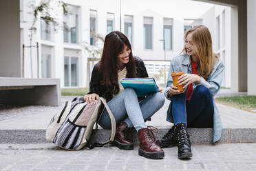 Fröhliche junge Studentinnen lachen, während sie auf den Stufen des Campus sitzen - TCEF01356