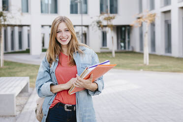 Lächelnde schöne blonde junge Frau hält Bücher auf dem Universitätscampus - TCEF01371