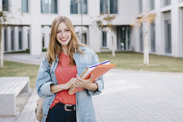 Smiling beautiful blond young woman holding books at university campus - TCEF01371