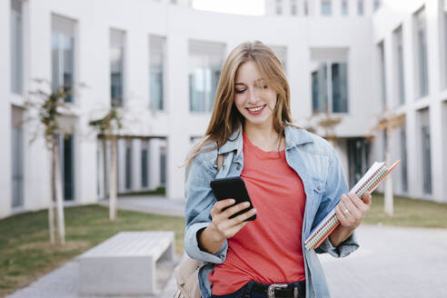 Smiling young blond female student using smart phone at university - TCEF01375