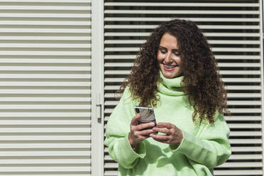 Lächelnde Frau im neongrünen Pullover, die ein Mobiltelefon vor einer Metalltür an einem sonnigen Tag benutzt - PNAF00242