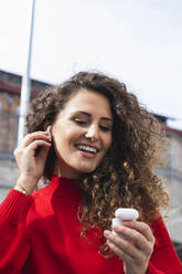 Happy woman connecting wireless in-ear headphones on sunny day - PNAF00255