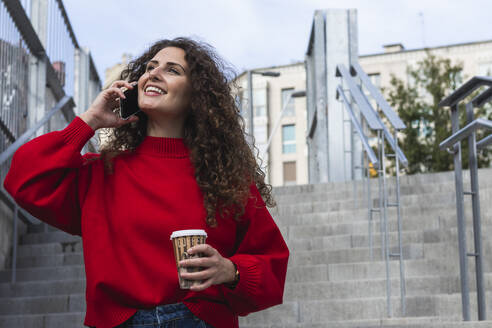 Lächelnde Frau, die eine Kaffeetasse hält, während sie auf einer Treppe mit einem Handy spricht - PNAF00262