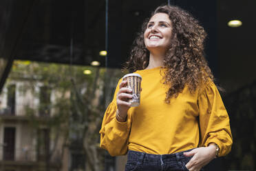 Happy businesswoman in yellow blouse holding disposable cup while looking up - PNAF00264