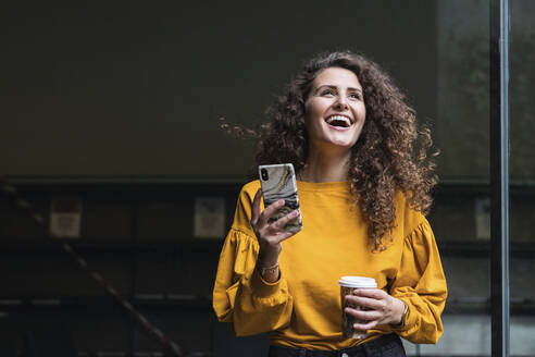 Fröhliche Frau mit lockigem Haar, die lachend ein Mobiltelefon und einen Einwegbecher hält - PNAF00266