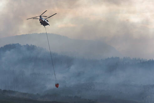 Militärhubschrauber fliegt mit Feuerschutzmittel, während Rauch von einem Waldbrand bei Sonnenuntergang aufsteigt - CAVF90814