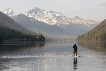 Rückenansicht eines anonymen Mannes auf einem Paddelbrett auf der ruhigen Oberfläche des Duffey-Sees und Genießen der Berglandschaft am Morgen in British Columbia - CAVF90872