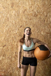 Schöne lächelnde junge Frau in Sportkleidung, die einen schweren Lederball hält und in die Kamera schaut, während sie in der Nähe einer Korkwand im Fitnessstudio steht - CAVF90875