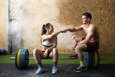 Starker Mann ohne Hemd und Frau in Sportkleidung, die lächeln und sich nett unterhalten, während sie im Fitnessstudio auf einer Langhantel sitzen - CAVF90879