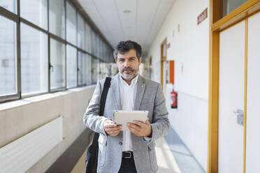 Älterer männlicher Professor, der ein digitales Tablet im Flur einer Universität hält - DGOF01720