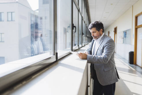 Professor, der ein digitales Tablet benutzt, steht auf dem Fensterbrett im Korridor einer Universität - DGOF01722