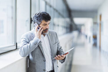 Männlicher Professor, der mit einem Mobiltelefon spricht, während er auf ein digitales Tablet am Fenster in einem Universitätskorridor schaut - DGOF01724