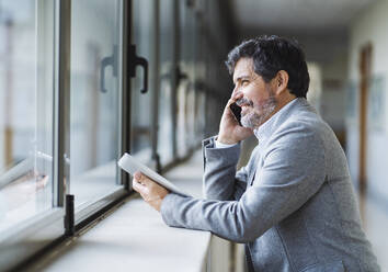 Lächelnder männlicher Professor, der am Telefon spricht und durch ein Fenster im Korridor einer Universität schaut - DGOF01726