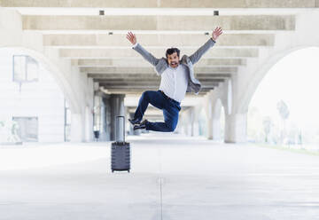 Happy male entrepreneur jumping with hands raised over concrete footpath - DGOF01731