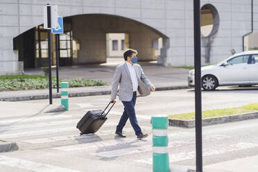 Geschäftsmann beim Überqueren der Straße mit Gepäck auf Rädern während der COVID-19 - DGOF01733