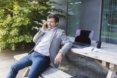 Smiling businessman talking through smart phone leaning on table against office building - DGOF01740