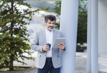 Männlicher Unternehmer, der ein digitales Tablet benutzt und eine Kaffeetasse in der Hand hält - DGOF01743