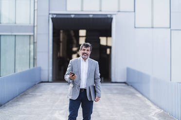 Smiling businessman with smart phone and headphones against office building - DGOF01747