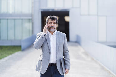 Male professional walking while talking on phone against office building - DGOF01749
