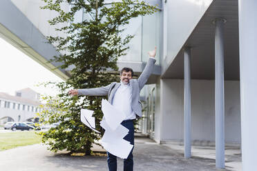 Cheerful businessman throwing documents while standing at office building - DGOF01751