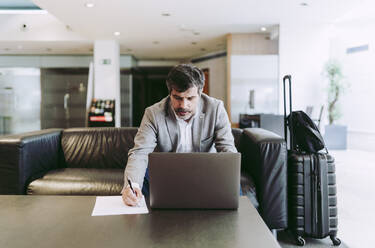 Älterer Geschäftsmann, der in der Hotellobby mit seinem Laptop auf Papier schreibt - DGOF01756