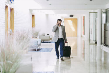 Male entrepreneur on phone pulling wheeled luggage in hotel corridor - DGOF01764