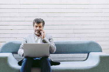 Businessman talking through mobile phone using laptop while sitting on sofa against wall in hotel - DGOF01766