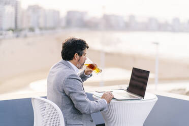 Male professional drinking beer while using laptop at cafe on hotel terrace - DGOF01781