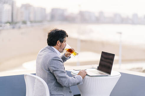 Männlicher Berufstätiger trinkt Bier und benutzt einen Laptop in einem Café auf einer Hotelterrasse - DGOF01781