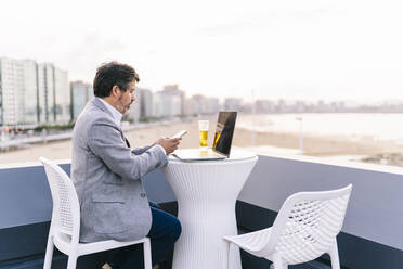 Männlicher Unternehmer, der ein Mobiltelefon benutzt, während er mit Bier und Laptop auf der Hotelterrasse sitzt - DGOF01782