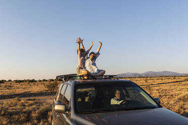 Teenage girl and her younger brother on top of SUV on desert road - MINF15422