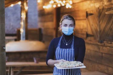 Woman waitress in apron and face mask holding fresh pizza on a board - MINF15447
