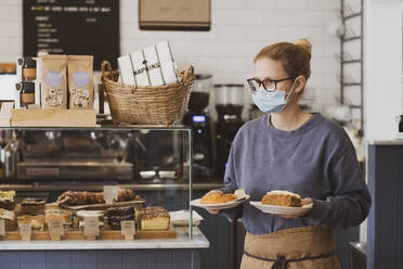 Kellnerin mit Gesichtsmaske, die in einem Café arbeitet und Teller mit Essen trägt. - MINF15450