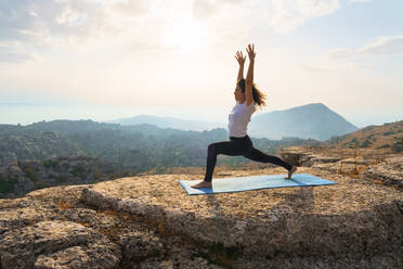 Full body of barefoot female standing in warrior pose on top of mountain while practicing yoga in nature at sunset time - ADSF17757
