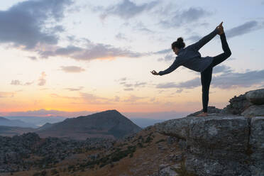 Niedriger Winkel Seitenansicht der flexiblen weiblichen stehend in Lord of the Dance Position gegen bewölkten Sonnenuntergang Himmel beim Üben Yoga auf der Spitze des felsigen Berg - ADSF17764