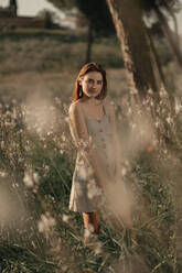 Portrait of young beautiful woman wearing a beige dress, posing in a field full of flowers and surrounded by trees. She is behind tall plants and backlit with sunlight at sunset. - ADSF17770