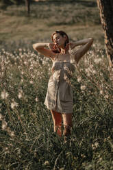 Porträt einer jungen schönen Frau in einem beigen Kleid, die in einem Feld voller Blumen und umgeben von Bäumen posiert, hinter hohen Pflanzen und im Gegenlicht der untergehenden Sonne. - ADSF17771