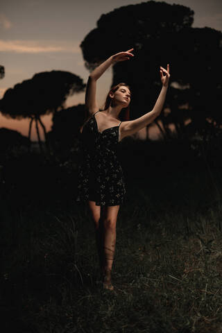 Elegant female ballet dancer in dress standing in meadow with raised arms and performing in dark park in Iceland stock photo