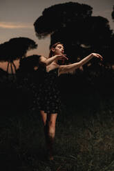 Elegant female ballet dancer in dress standing in meadow with raised arms and performing in dark park in Iceland - ADSF17776