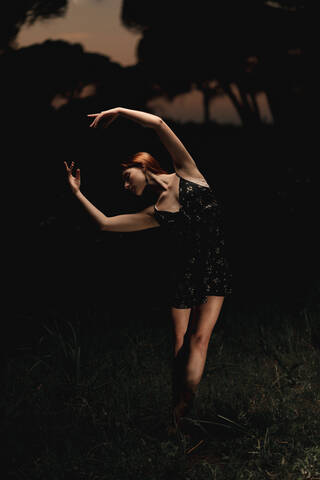 Elegant female ballet dancer in dress standing in meadow with raised arms and performing in dark park in Iceland stock photo