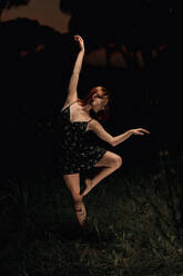 Elegant female ballet dancer in dress standing in meadow with raised arms and performing in dark park in Iceland - ADSF17778