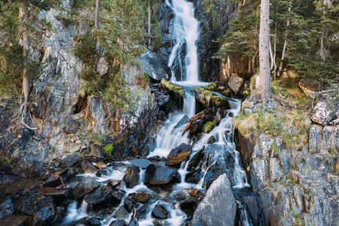 Langer schmaler Wasserfall eines Gebirgsflusses, der über große Steine zwischen Bäumen auf Felsen in einer Schlucht fließt - ADSF17788