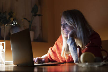 Lächelnde Frau, die einen Videoanruf auf dem Laptop hört, während sie zu Hause am Tisch sitzt - ERRF04695
