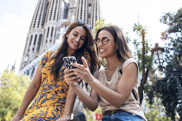 Frauen, die ein Mobiltelefon benutzen, während sie vor der Sagrada Familia in Barcelona, Katalonien, Spanien sitzen - JSRF01173